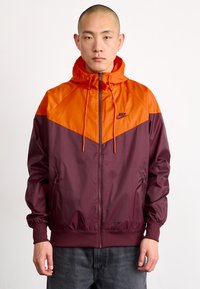 Chaqueta Nike con capucha, que presenta un diseño en bloques de color naranja y burdeos. Fabricada con una tela suave y ligera, con puños elásticos y bolsillos con cremallera.