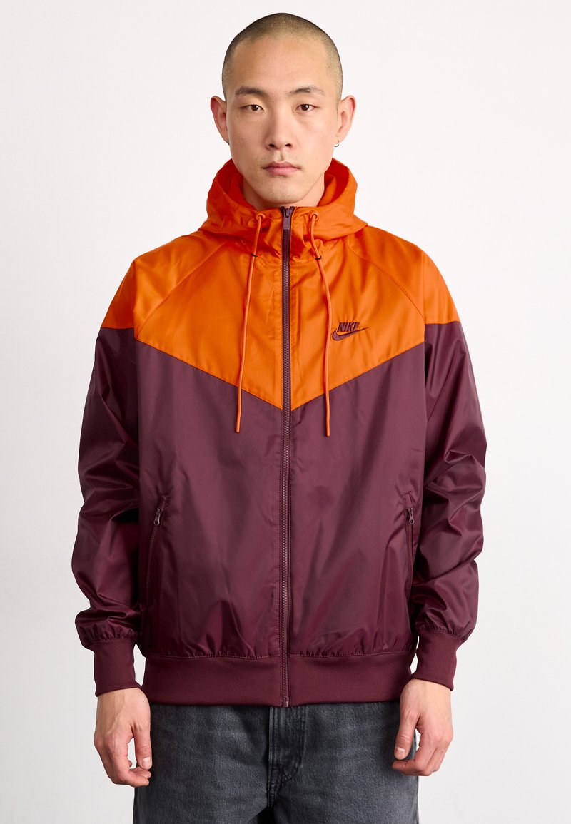 Chaqueta Nike con capucha, que presenta un diseño en bloques de color naranja y burdeos. Fabricada con una tela suave y ligera, con puños elásticos y bolsillos con cremallera.