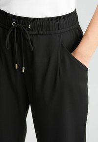 Schwarze Jogginghose mit elastischem Bund, Kordelzugverschluss, Seitentaschen und goldfarbenen Hardware-Akzenten. Glatte Stofftextur.