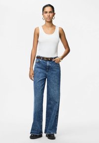 Femme portant un haut blanc sans manches, un jean bleu taille haute à jambes larges avec une ceinture marron, des chaussures noires et des boucles d'oreilles créoles dorées.