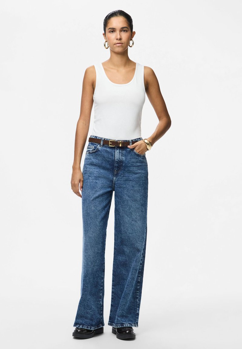 Femme portant un haut blanc sans manches, un jean bleu taille haute à jambes larges avec une ceinture marron, des chaussures noires et des boucles d'oreilles créoles dorées.