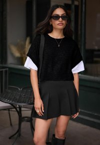Zwarte, cropped gebreide trui over een wit shirt, gecombineerd met een geplooide zwarte minirok. Accessoires zijn onder andere gouden ringen en een zonnebril.