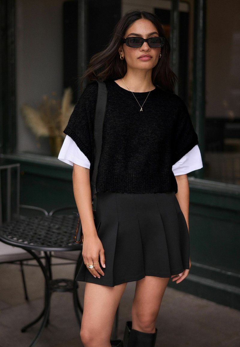 Zwarte, cropped gebreide trui over een wit shirt, gecombineerd met een geplooide zwarte minirok. Accessoires zijn onder andere gouden ringen en een zonnebril.