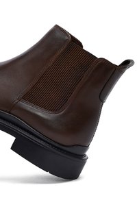 Stivale Chelsea in pelle marrone con pannello laterale elasticizzato, linguetta e suola nera spessa su fondo bianco.