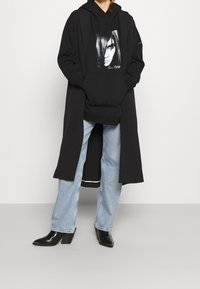 Henkilö, jolla on päällään musta pitkä takki sekä musta painokuvioitu huppari, vaaleansiniset farkut ja mustat korolliset saappaat, seisomassa yksiväristä taustaa vasten.