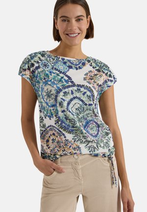 Femme souriante portant un haut blanc à manches courtes avec des motifs circulaires bleus, verts et orange et un pantalon beige avec la main dans la poche.
