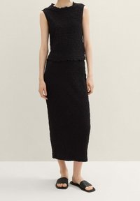 Černý texturovaný dvoudílný outfit obsahující top bez rukávů se zvlněným lemem a odpovídající dlouhou sukni, doplněný černými skluznými sandály.