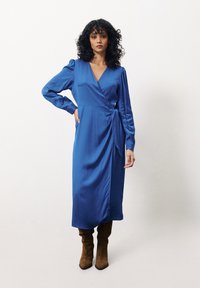 Blaues Wickelkleid mit V-Ausschnitt, langen Ärmeln und seitlichem Bindeband. Glatte Satin-Textur, knielang, kombiniert mit braunen Stiefeln bis zum Knie.