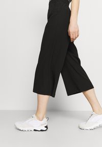 Black wide-leg culotte pants with an elastic waistband, featuring a smooth texture. Paired with white athletic shoes.