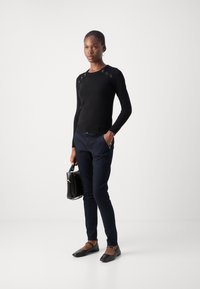 Černý pletený svetr s ozdobami na ramenech, spárovaný s tmavě modrými úzkými kalhotami a černými balerínami. Modelka drží černou kabelku.