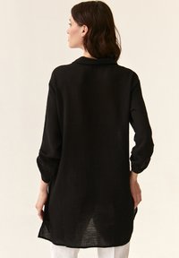 Black, long-sleeve tunic made from textured fabric. Features a relaxed fit, curved hem, and a collar. No visible fasteners or patterns.