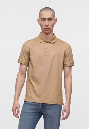 Alvorlig ung mand iført en beige polo med korte ærmer og blå jeans, stående foran en ensfarvet hvid baggrund.
