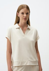 Helles beiges Poloshirt mit V-Ausschnitt, kurzen gerollten Ärmeln und lockerer Passform. Aus weichem, glattem Stoff mit einer subtilen Textur gefertigt.