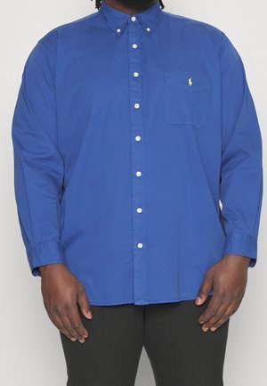 Homme portant une chemise bleue à manches longues avec un petit logo jaune brodé sur la poche poitrine.