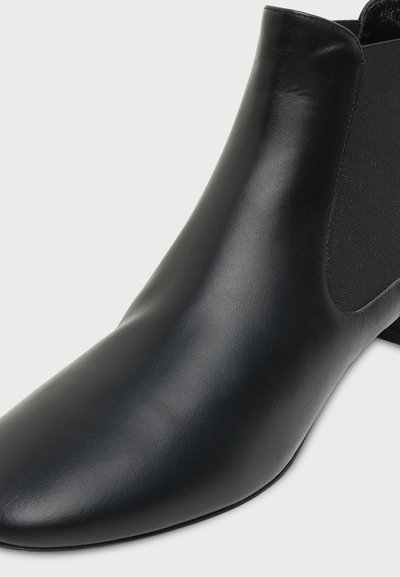Gros plan d'une botte cheville en cuir noir avec un panneau latéral élastique et un talon bas sur un fond clair.