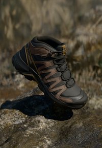 Tamsiai pilka ir ruda „Salomon“ žygiavimo batai su „Gore-Tex“, raišteliais ir tekstūruotu padu, ilsintis ant akmenuoto paviršiaus.