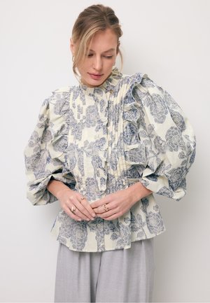 Vrouw die een crèmekleurige blouse met blauwe bloemmotieven en ballonmouwen draagt, gecombineerd met lichtgrijze broek met wijde pijpen, terwijl ze naar beneden kijkt.
