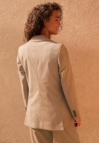 Light gray suit jacket with a tailored fit, notch lapels, and buttoned cuffs. Smooth texture with subtle sheen against a beige background.