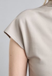 Lichtbeige, gestructureerde stoftop met een relaxte pasvorm. Korte mouwen met een subtiele drop-shoulder ontwerp en nette stikdetails.