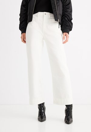 Άτομο που φοράει λευκό παντελόνι με φαρδιά μπάσκα, μαύρες γυαλιστερές μυτερές μπότες και μαύρο μπουφάν τύπου bomber με φερμουάρ.