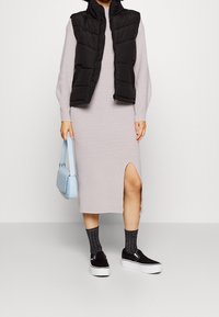 Světle šedé dlouhé šaty s rozparkem na boku, doplněné černou péřovou vestou, šedými ponožkami s vzorem a černými teniskami na nazutí. Světle modrá kabelka.