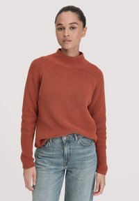 Rippiger rostfarbener Pullover mit hohem Kragen und langen Ärmeln, kombiniert mit hellblauen geraden Jeans. Die gemütliche Stricktextur ist deutlich erkennbar.