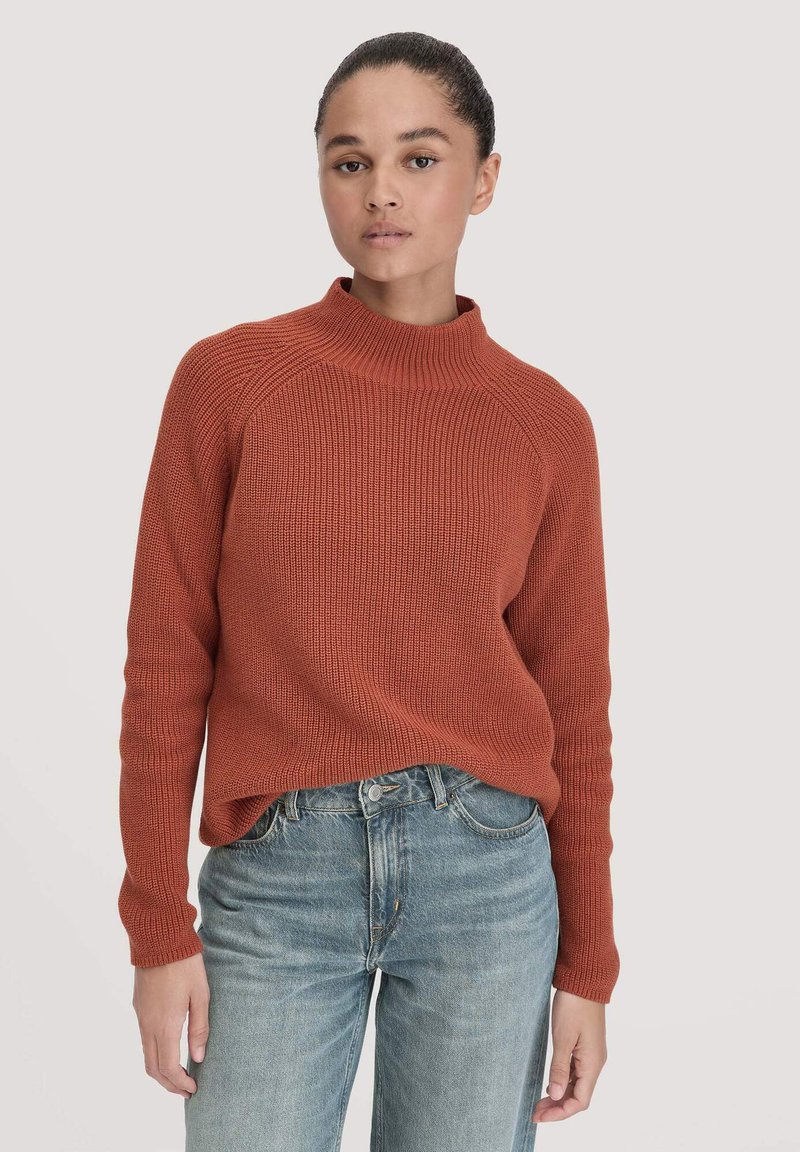 Rippiger rostfarbener Pullover mit hohem Kragen und langen Ärmeln, kombiniert mit hellblauen geraden Jeans. Die gemütliche Stricktextur ist deutlich erkennbar.