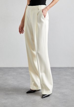 Λευκά παντελόνια με φαρδιά γραμμή από λείο ύφασμα, με εμπρός πτυχώσεις, πλαϊνές τσέπες και ραμμένο λάστιχο στη μέση. Συνδυάζονται με μαύρα μυτερά παπούτσια.