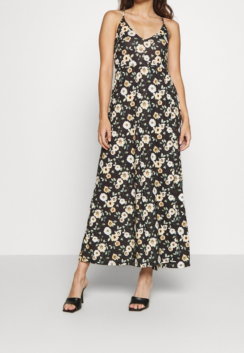 Femme portant une longue robe noire sans manches à motifs floraux jaunes et blancs, sandales à talons noires, debout devant un fond uni.