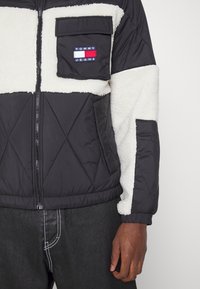 Pěnová černá bunda s krémovými fleecovými sekcemi, obsahující náprsní kapsu s logem, zapínání na zip a diagonální prošívaný vzor na těle.