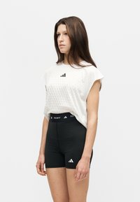 Femme debout, tournée vers la gauche, portant un haut sans manches blanc texturé et un short de sport noir taille haute avec des logos de marques.
