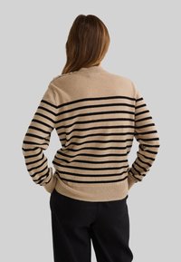 Personne aux cheveux longs bruns, portant un pull beige à rayures noires horizontales et un pantalon noir, se tenant les mains sur les hanches.