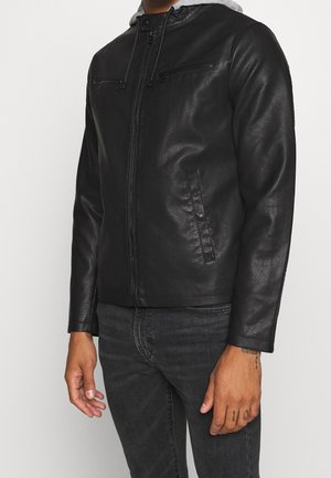 Mężczyzna ubrany w czarną skórzaną kurtkę z zamkiem błyskawicznym i kieszeniami na piersi na zamek, w połączeniu z ciemnymi dżinsami na jednolitym białym tle.