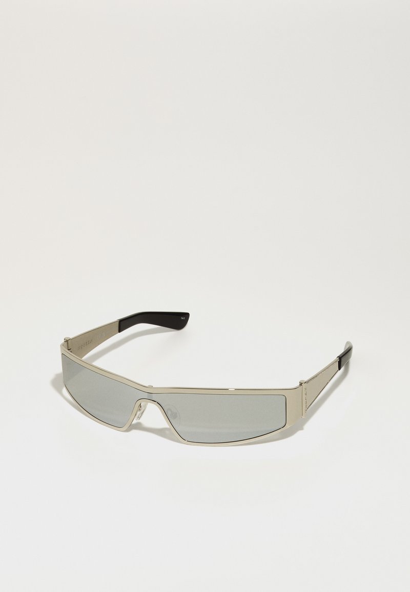 Gafas de sol con montura metálica plateada y lentes ahumados grises. El diseño es angular, con brazos delgados que tienen puntas negras y acentos texturizados.