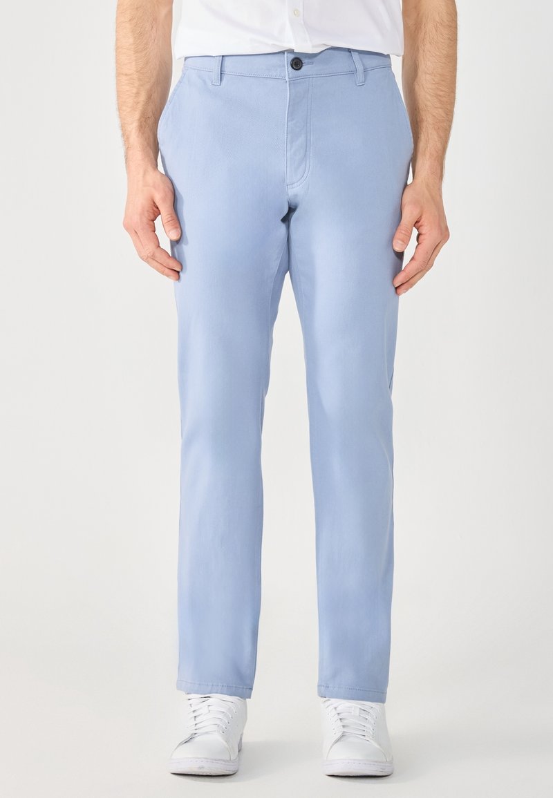 Homme portant un pantalon slim bleu clair, des baskets blanches et une chemise blanche rentrée, debout devant un fond uni.