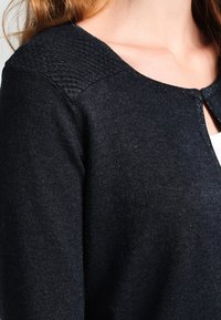 Cardigan de punto negro con cuello redondeado y detalles texturizados en los hombros. Tejido suave, con cierre de botón en la parte delantera.