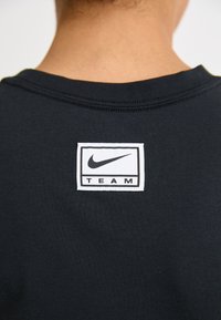 Črna majica s kratkimi rokavi z belim pravokotnim Nike oznako na zadnjem ovratniku, pod logotipom pa je napis "TEAM". Gladka tekstura blaga.