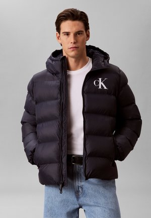 Giovane uomo con giacca imbottita nera con cappuccio, t-shirt bianca e jeans azzurri, in piedi su sfondo grigio.
