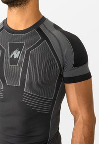 Približnji posnetek moškega, ki nosi črno-sivo prilegajočo športno majico z teksturiranimi geometričnimi vzorci in majhnim logotipom gorile na prsih.