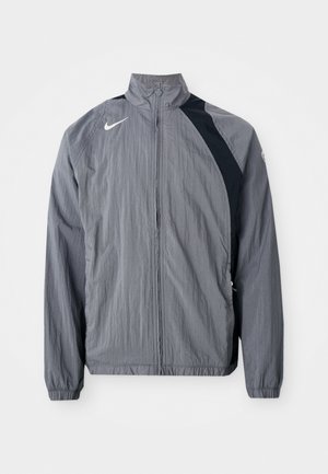 Grå letvægtsjakke lavet af tekstureret stof, med høj krave, lynlås lukning og sorte accenter på den ene side. Nike-logo synligt.