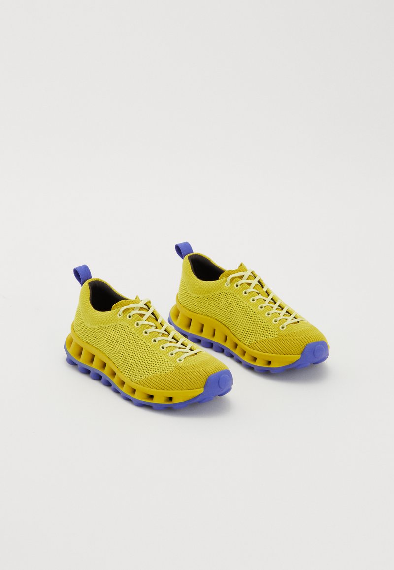 Gelbe Stricksneaker mit lila Sohlen und Fersenlaschen, ausgestattet mit weißen Schnürsenkeln und einer belüfteten Dämpfungsmittelsohle.