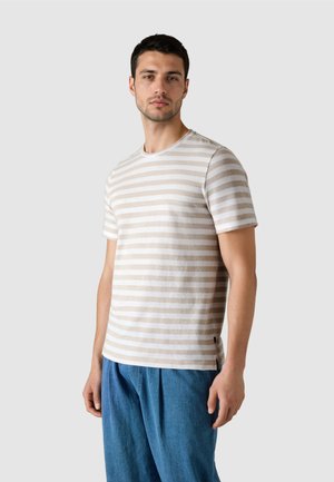 Hombre vistiendo una camiseta de manga corta a rayas beige y blanca y pantalones azules sueltos, de pie frente a un fondo gris liso.