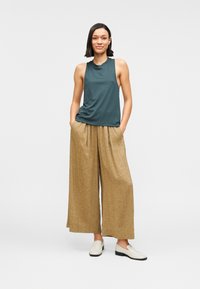 Top sin mangas color verde azulado combinado con pantalones anchos de pierna ancha y tono marrón claro. Zapatos blancos tipo slip-on con textura suave y detalles plisados en los pantalones.