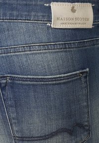 Jeans in denim con una texture blu sbiadita, caratterizzati da una tasca posteriore e un'etichetta in pelle con la scritta "MAISON SCOTCH." Cuciture di un blu più scuro.