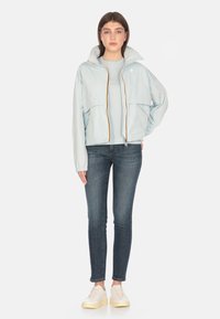 Giacca impermeabile azzurro con zip, dotata di tasche oversize e cappuccio, abbinata a un top azzurro, jeans skinny scuri e sneakers bianche.