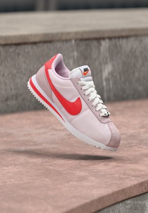 Svijetloružičasta i crvena Nike tenisica s bijelim vezicama, suspendirana u zraku iznad kamene površine.