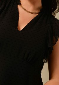 Musta mekko, jossa on V-pääntie, lyhyet röyhelöhiha, pistekuvioitu tekstuuri ja kerätty vyötärö. Näkyvissä kultaketju-kaulaketju.