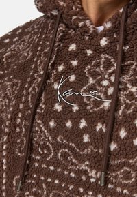 Brown fleece hoodie with a textured surface, featuring a white pattern, drawstrings, and metallic logo detail on the front.