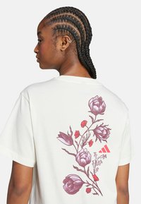 Camiseta de algodón color crema con un gráfico floral en rosa y rojo en la parte de atrás, con un pequeño acento de logo rojo. Mangas cortas.