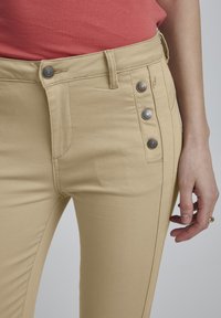 Bež hlače od pamuka s mješavinom vlakana, glatke teksture, slim fit kroja, s pet dugmadi sprijeda i dekorativnim šavovima sa strane.